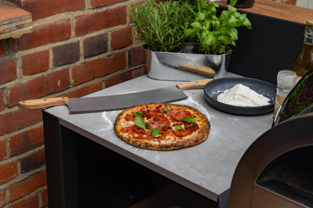 pizza on worktop next to pizza oven with pizza cutter