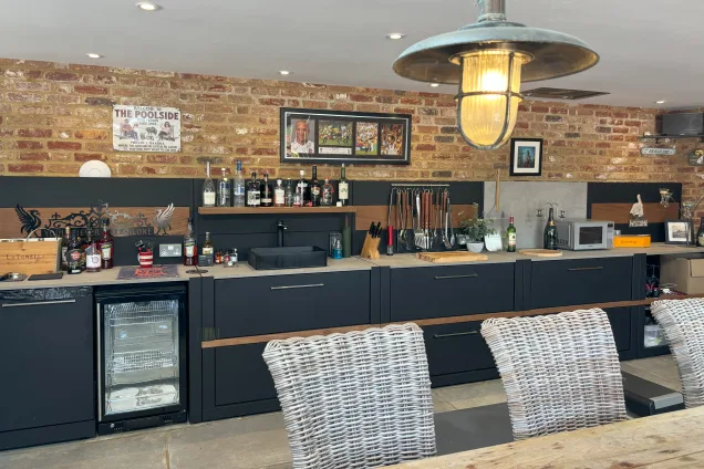 Outdoor kitchen in saffron walden with storage units and a drinks fridge with lots of bottles of drink on the feature wall shelf and the worktop
