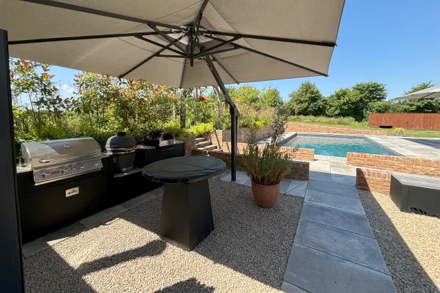 Outdoor kitchen in saffron walden under canopy with chefs anvil, gas bbq, charcoal bbq and pizza oven on patio with pool in background