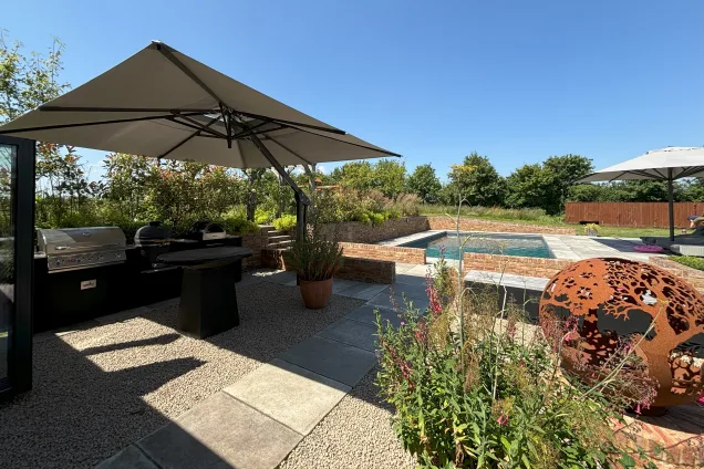Outdoor kitchen in saffron walden under canopy with chefs anvil, gas bbq, charcoal bbq and pizza oven on patio with pool and flowers in background