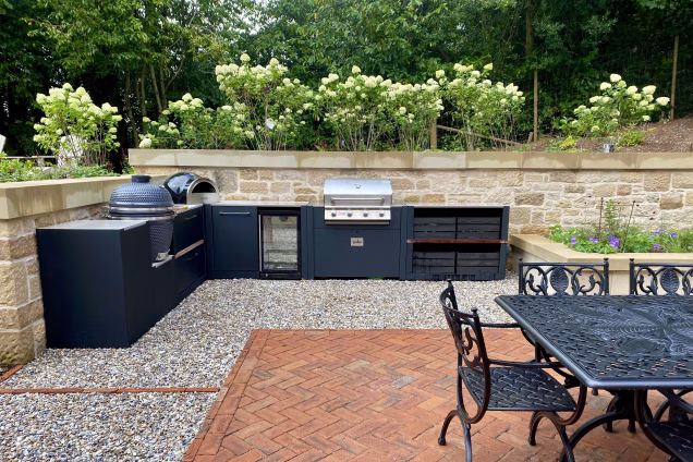 outdoor kitchen in front of stone wall on stones with gusto charcoal bbq, pizza oven and bull gas grill and storage cabinet and bin and fridge