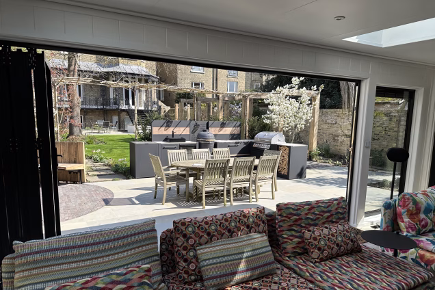 View looking out of a garden building with a Grillo kitchen in the centre of the frame. Oak framing surrounds a garden path and a Victorian style wrought iron lead-roofed verandah is on the house in the background