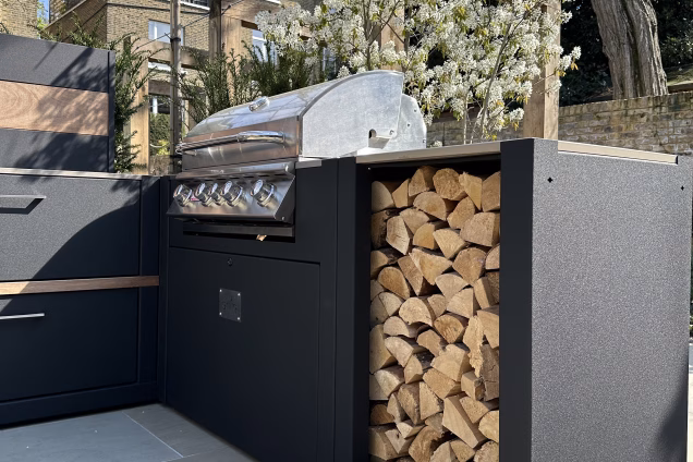 Closeup photo of Bull gas grill and Grillo Tall Square corner module filled with logs, with a blossoming Serviceberry tree in the background