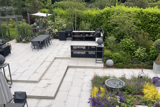 A photo from an upstairs window looking out across a residential garden in Cranleigh, Surrey. A Grillo outdoor kitchen and dining area can be seen, along with well-tended flowering borders