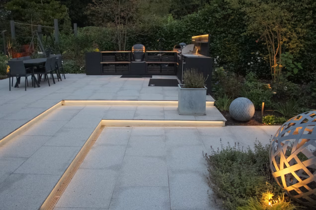 A wide angle shot showing custom garden lighting alongside paths, under steps and integrated into the outdoor kitchen in a residential garden in Cranleigh, UK