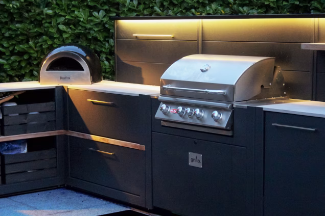 A closeup view of a Bull gas grill and Delivita pizza oven in an illuminated Grillo outdoor kitchen at dusk