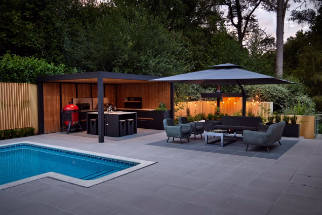 Grillo outdoor kitchen and comfy seating area under a pergola and parasol respectively, shown at dusk with atmospheric lighting