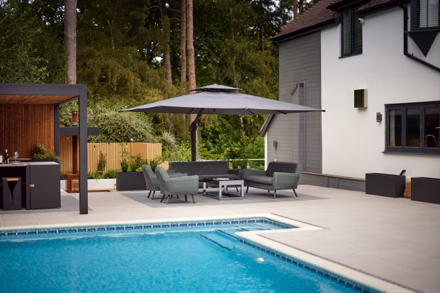 Photo looking over a swimming pool towards a parasol seating area, along the side of a large house in Ashtead, Surrey