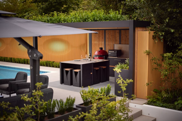 Telephoto image looking towards a Grillo outdoor kitchen against a modern orange garden wall, with a parasol and hedging in the foreground