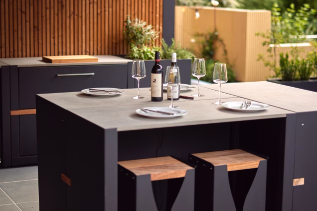 Photo of an outdoor kitchen bar table with seating for six. The image is focused on the wine bottles and glasses on the worksurface, with the rest of the image gently blurred