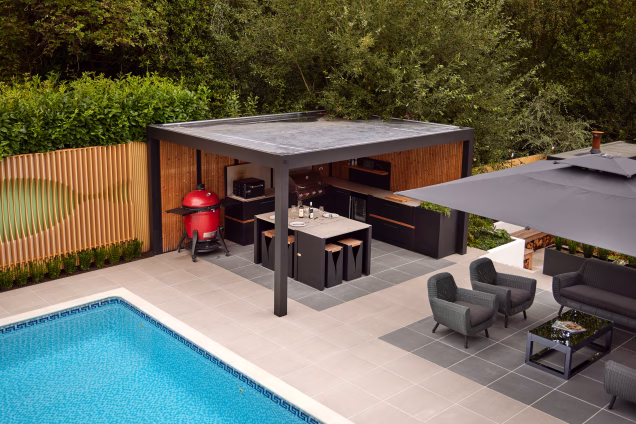 Photo from the upstairs window of a house overlooking the side garden, where an outdoor kitchen by Grillo can be seen under a pergola, with dense vegetation beyond