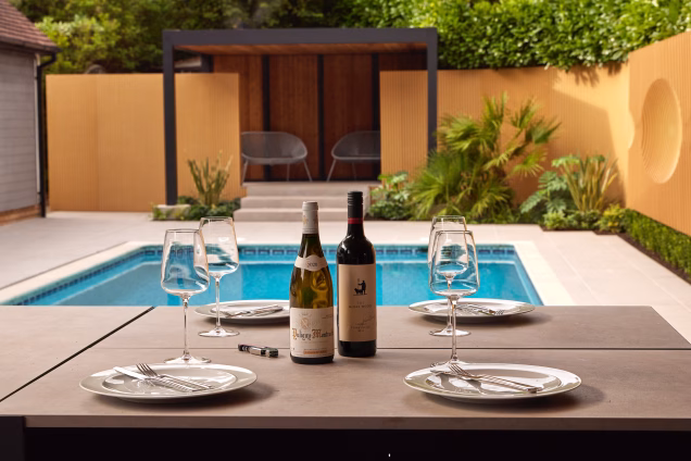 Lifestyle photo of wine bottles and glasses on a laid outdoor kitchen bar table, overlooking a pool and seating area in the background