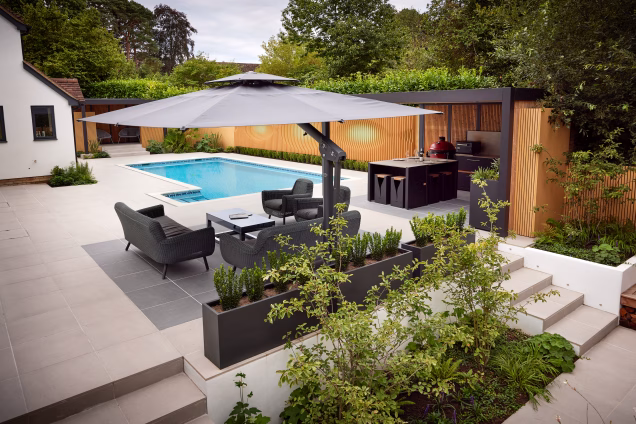 Photo looking along a side garden with a large parasol and seating area in the foreground and a Grillo outdoor kitchen and swimming pool in the background