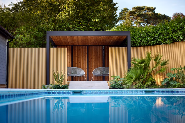 Low level photo looking along the surface of a swimming pool towards two garden chairs under a pergola overlooking the pool