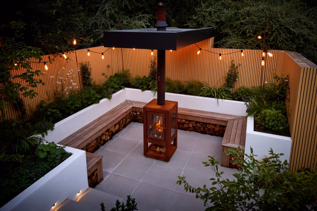 A suspended firepit area in lower garden, subtly lit at dusk, and lined with plants, surrounded by warm-toned vertical narrow-slat fencing