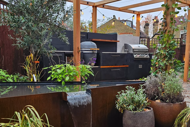 Looking over a plant-fringed water feature to a Grillo outdoor kitchen in a garden in Dulwich, London.