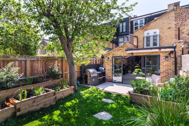 A wide-angle view of the garden of a property in Stoke Newington, with stepping stones on the grass leading the eye into the centre of the image, where there's an outdoor kitchen on the patio