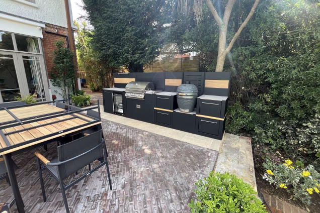 A wide angle view of an outdoor kitchen in Richmond set amongst subtropical plants, such as a New Zealand palm and "Brachyglottis greyi" shrubs with beautiful yellow daisy-like flowers