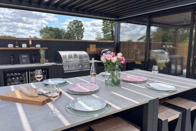 A bar table with laid places for 4, glasses of rose wine and a vase of roses under a pergola with an outdoor kitchen