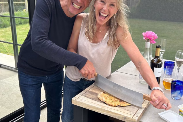 A photo of Sara and her husband in their outdoor kitchen laughing as they cut a pizza together