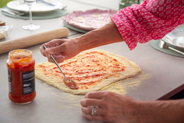 Closeup of hands holding a spoon spreading passata over a rectangular pizza base