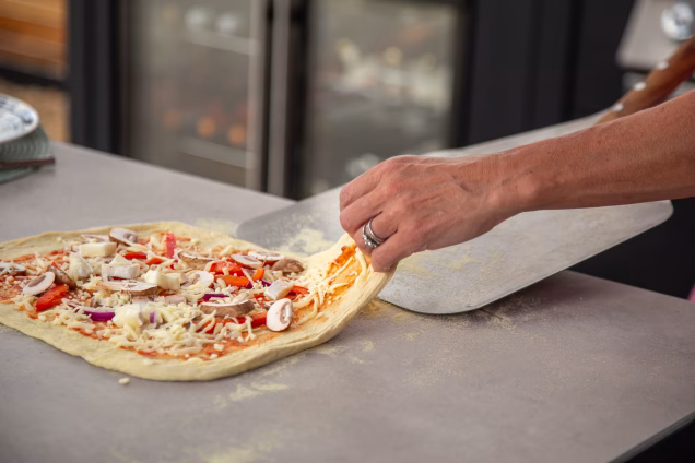 A raw pizza is lifted before the peel is slid under