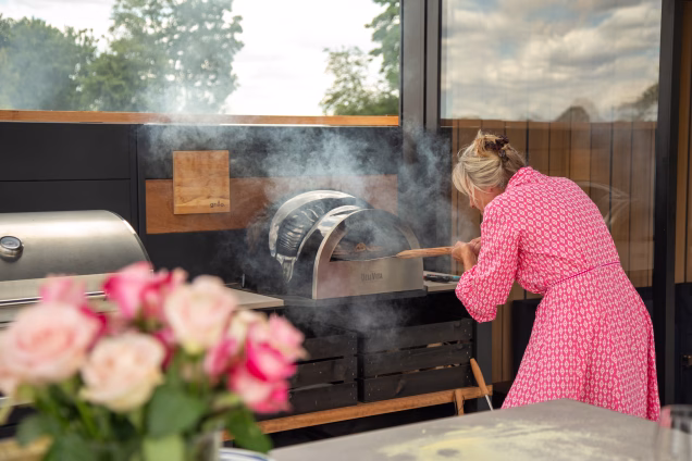 A woman in a pink dress turns a pizza in a smoking Delivita pizza oven in an outdoor kitchen