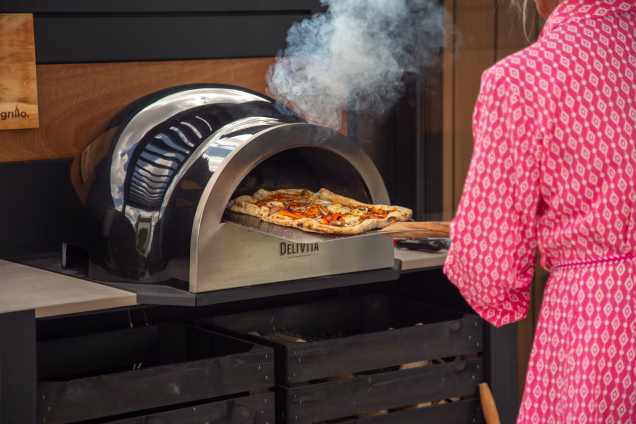 Closeup photo of a woman in a pink dress removing a rectangular pizza from a Delivita pizza oven in a Grillo outdoor kitchen