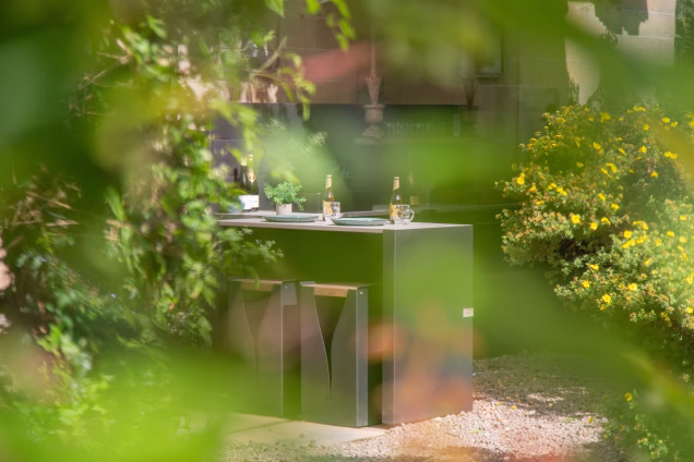 Looking through a blur of green foliage in the foreground to the bar and bar seating of a Grillo kitchen at a Baronial manor in the Scottish Borders, by the River Tweed