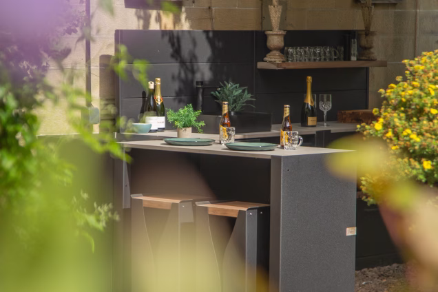 Looking through blurred foliage and purple flowers at the bar seating area of an outdoor kitchen in the Scottish Borders