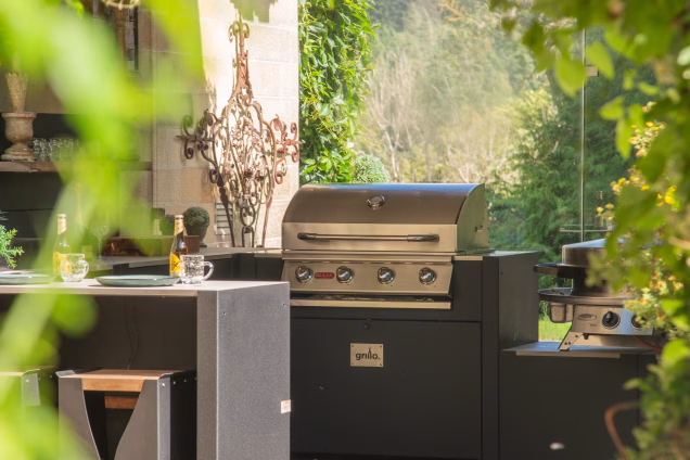 A photo with out-of-focus leaves bordering the frame, with the subject being the Bull gas grill in a Grillo kitchen in a sandstone archway at a castle in the Scottish Borders