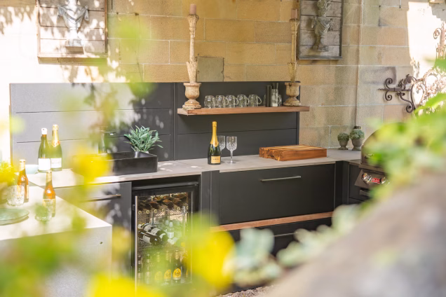 Looking through blurred leaves and yellow flowers towards the fridge and sink in a Grillo outdoor kitchen in the Scottish borders, with bright sunshine