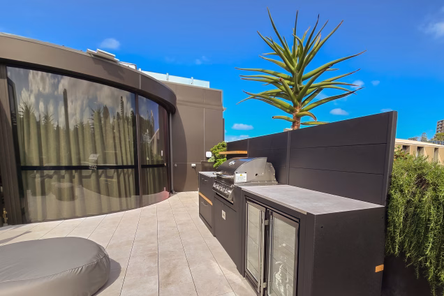 Looking along a roof terrace outdoor kitchen with the curved glass facade of a penthouse-level office in the background