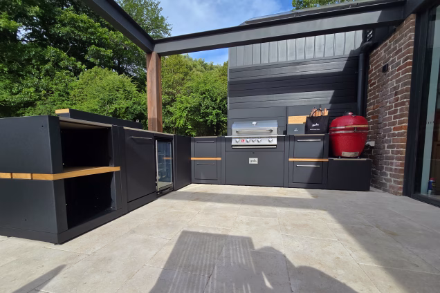 An outdoor kitchen featuring a red kamado BBQ, a powerful Bull Angus gas grill, a Vantage Drinks Station, fridge and standing bar
