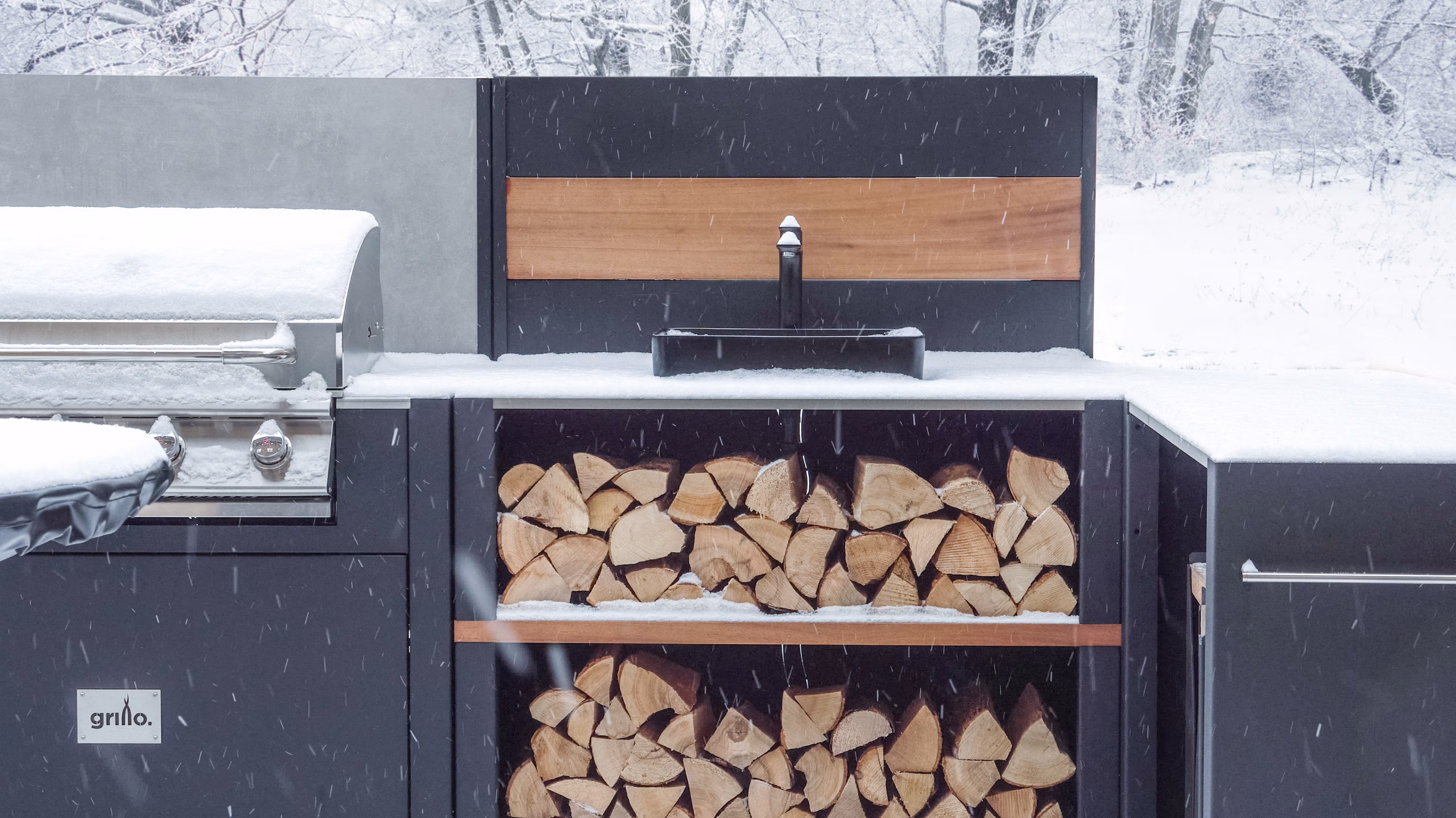 Grillo Vantage outdoor kitchen in heavy snow, with a gas grill to one side and a sink module centre of photograph, and snowy logs on the shelf underneath